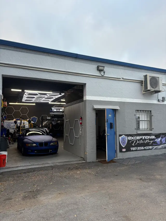 The Exceptional Detailing bay with its roll-up door open, a blue BMW Z3 under the hex light ceiling, the shop banner by the walk-in door